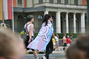 Two people walking along a street at a pride event. One has a trans pride flag draped over their shoulders. The other has rainbow braces holding up their shorts.