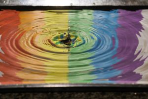 A drop falling into water with ripples spreading out across the surface. There are reflections of a rainbow pride flag.