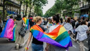 Looking along a street full of pride marchers. In the foreground are two people, holding hands, with a pride flag draped over the shoulders of both of them.