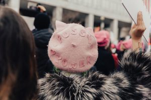 Head and shoulders image of the back of someone at a protest. They are wearing a pastel pink hat with the words "pussy power" on it.