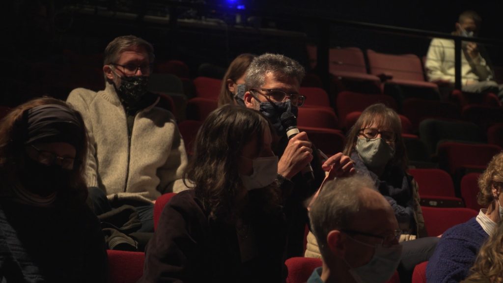 People who are sat in the audience wearing masks are interacting with the actors. One of the audience members is speaking into a microphone that they've been given.