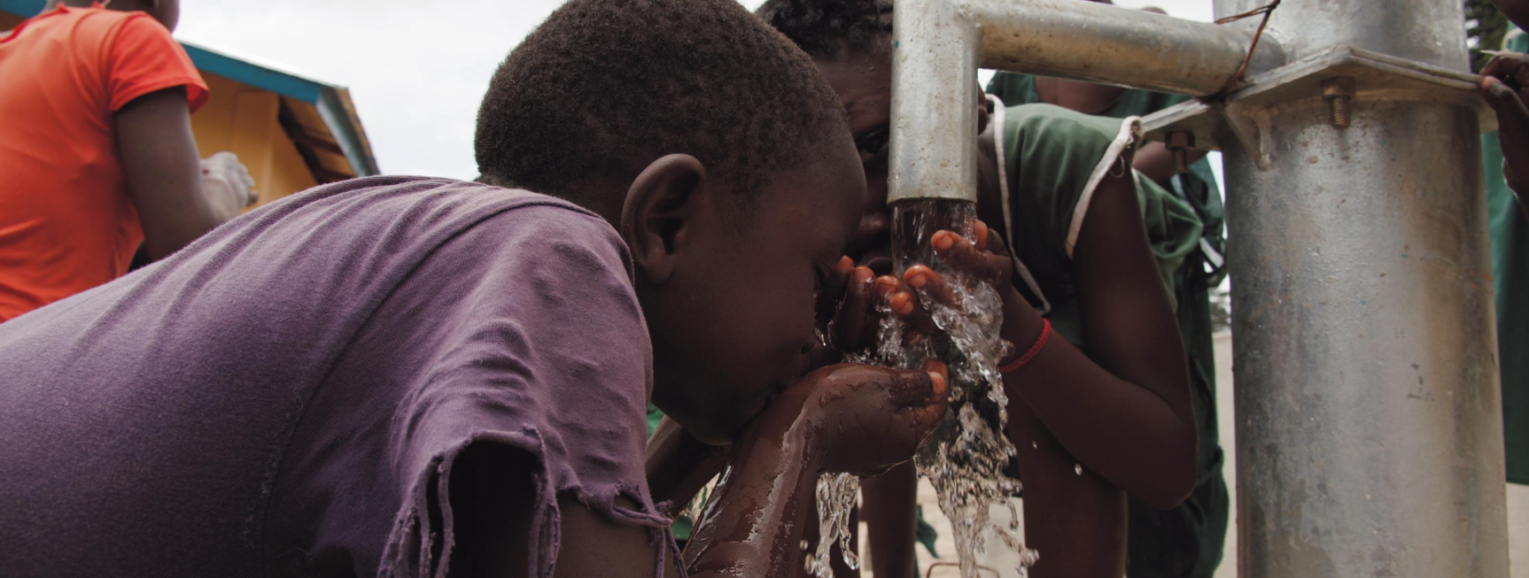 An image of an African child drinking clean water from a water pump. The child is cupping their hands together and bringing the water to her mouth.