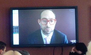 Image of Glen Cohen, a man with red framed glasses presenting to the audience via Skype.