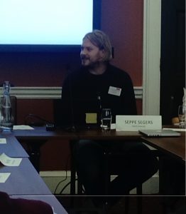 Image of Seppe Segers, a man dressed in black sitting behind a desk in front of a screen displaying a presentation.