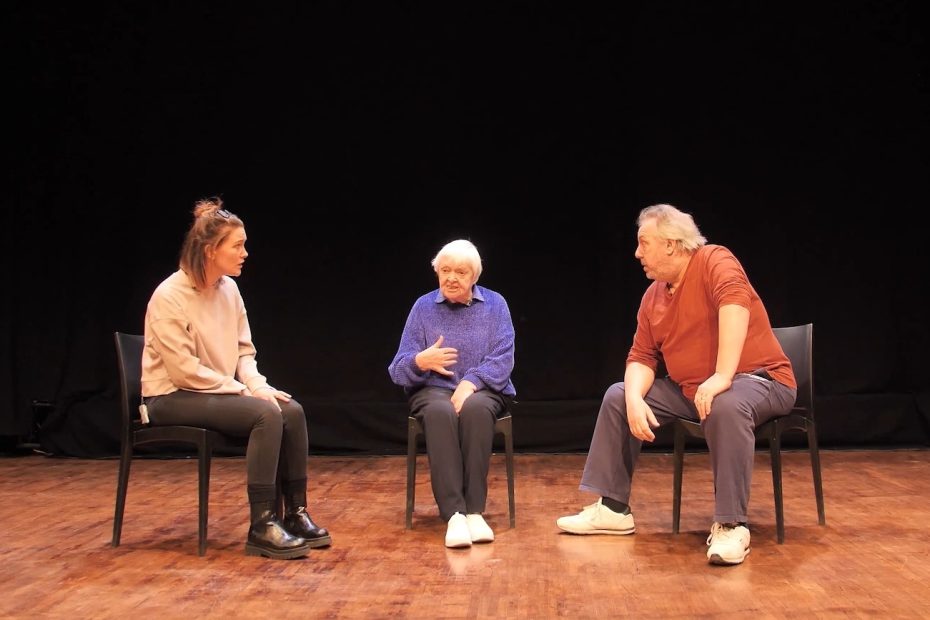 Three people sit on chairs onstage. The oldest person in the middle talks while the other two people listen.