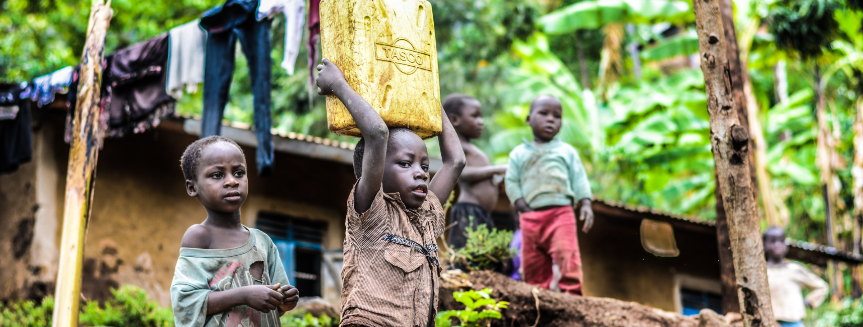 An image of a child carrying a bucket of water above their head. The child appears to live in poverty and in stood infront of a 'slum' with several friends.